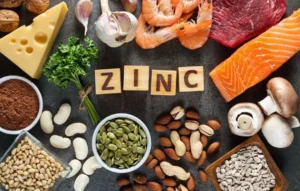Foods high in zinc such as cheese, shrimp, beef, salmon, garlic, parsley, mushrooms, almonds, peanuts, pumpkin seeds, sunflower seeds, beans, pine nuts, and cocoa powder, with wooden blocks spelling “ZINC.