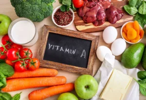 Assorted foods rich in vitamin A including carrots, broccoli, tomatoes, spinach, liver, eggs, milk, green apples, avocado, dried apricots, and butter, with a chalkboard labeled “Vitamin A.
