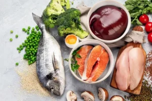 Fresh fish, salmon fillets, chicken breast, broccoli, green peas, mushrooms, leafy greens, egg yolk, liver, and grains arranged on a table.